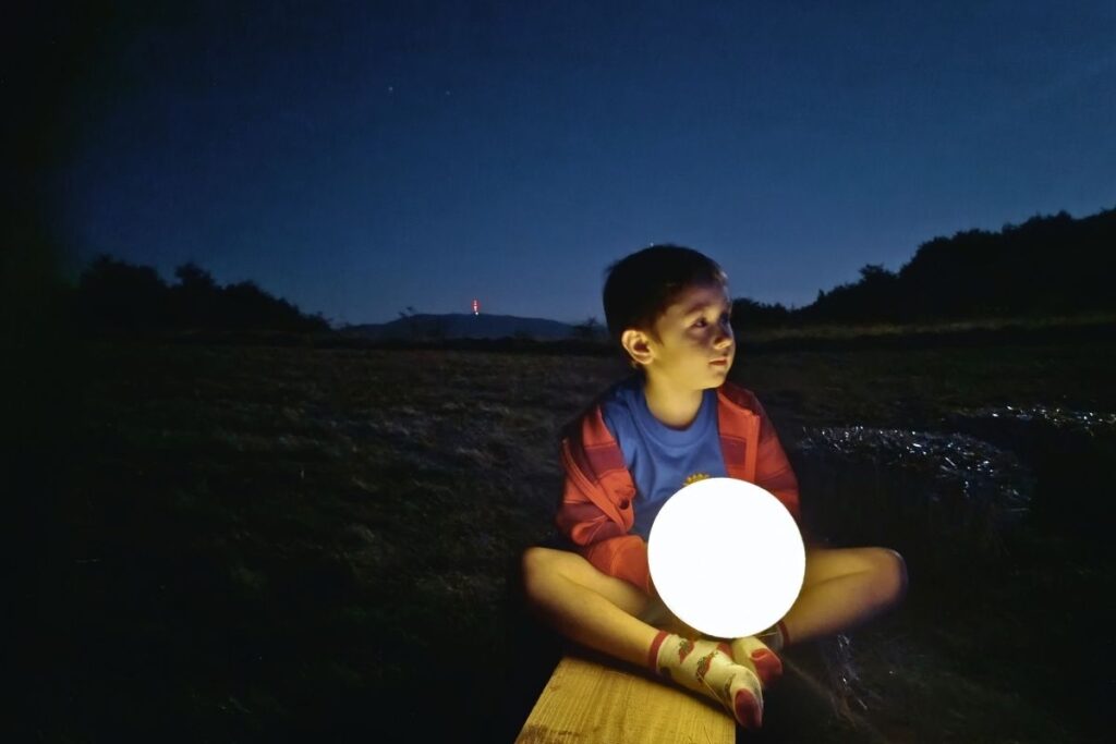 Dečak sedi na balama sena držeći svetleću kuglu, noćna scena pod zvezdanim nebom na planini Slemen